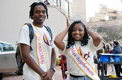 A male and a female student on the 亚洲博彩网站 plaza. They are wearing royal sashes.